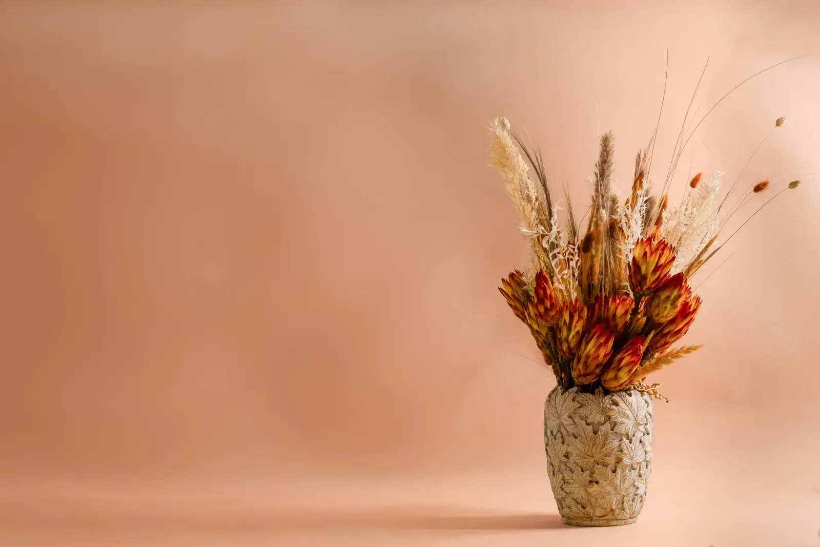 Trockenblumenstrauß mit orangen Proteas und Gräsern in einer Keramikvase vor apricotfarbenem Hintergrund