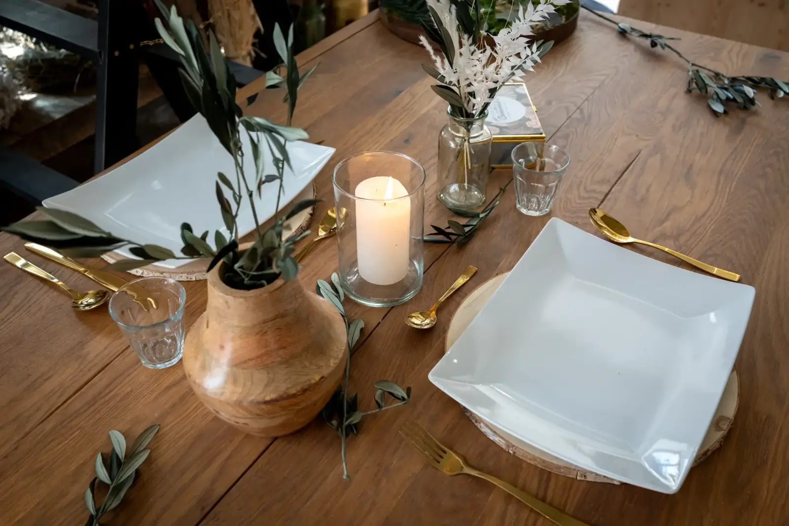 Gedeckter Holztisch mit Holzvase, Olivenzweigen, Kerze und weißen Tellern